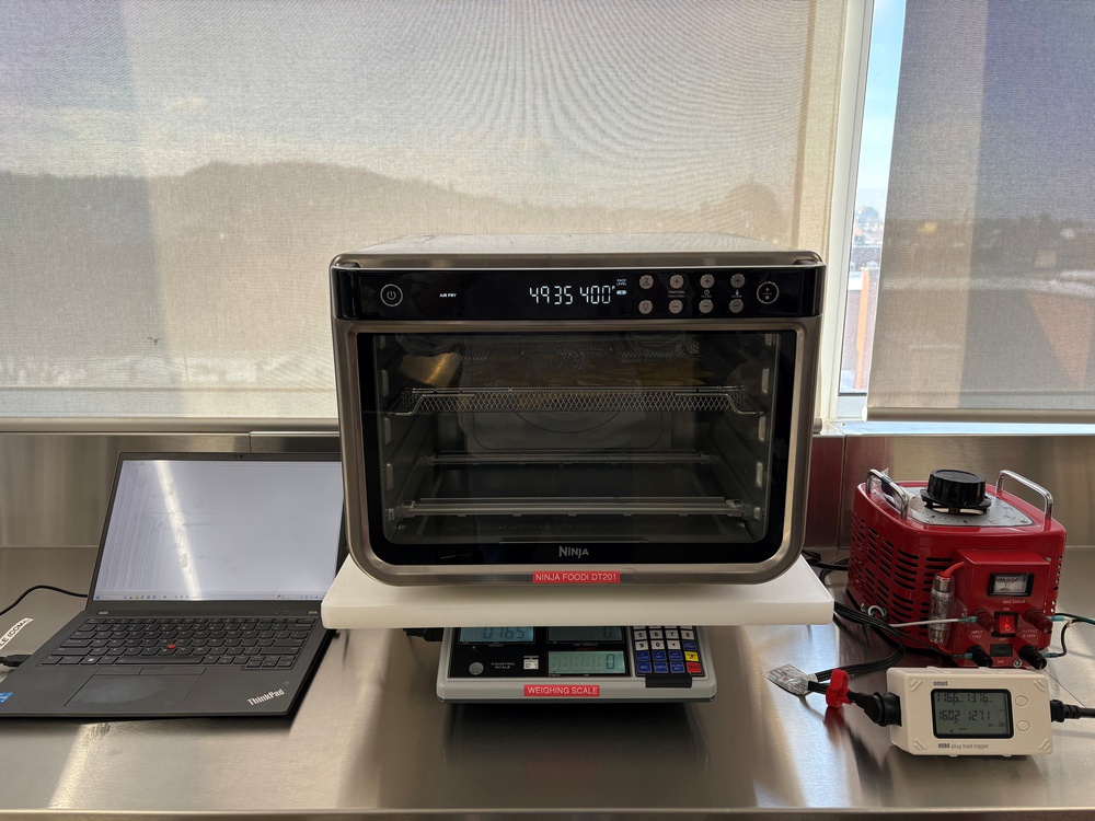 Testing setup for a Ninja Foodi DT201 oven, with a laptop, weighing scale, power monitor, and variable transformer on a stainless steel countertop,