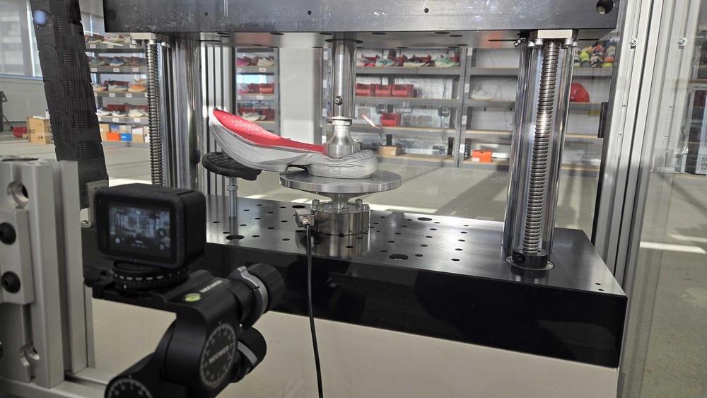 A heel compression test being conducted on the adidas Adizero Adios Pro 4 using a universal testing machine, with a cut shoe placed between the test head and base plate. A camera is set up nearby to record the process.